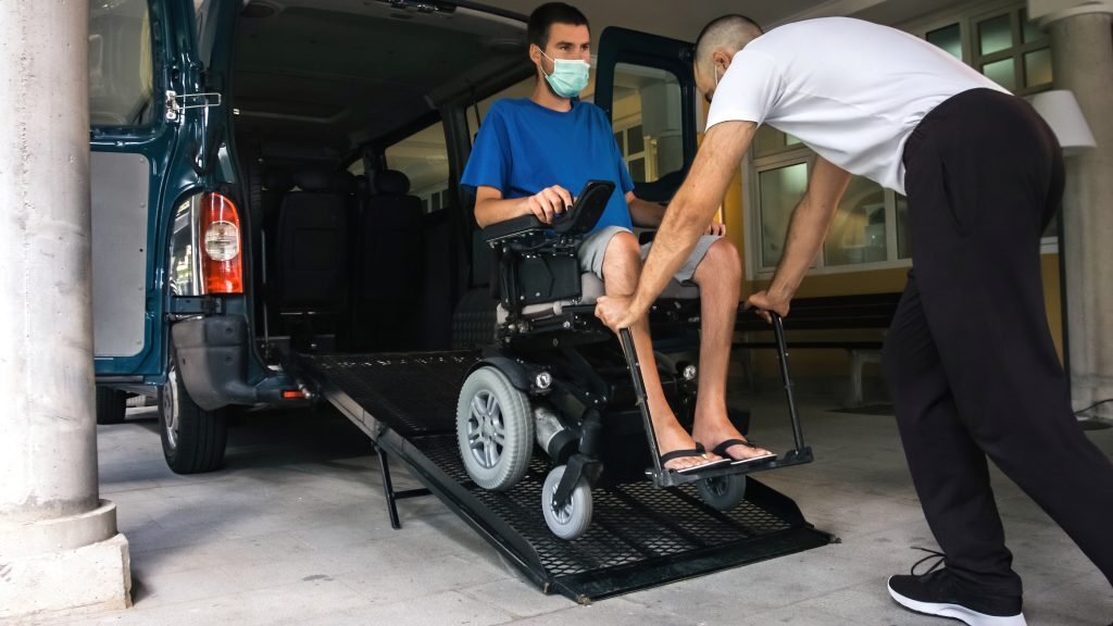 A driver helps a man who utilizes a wheelchair into his accessible vehicle. When patients use NEMT services to attend regular medical care instead of the ambulance to emergency services, emergency room diversion is successful.