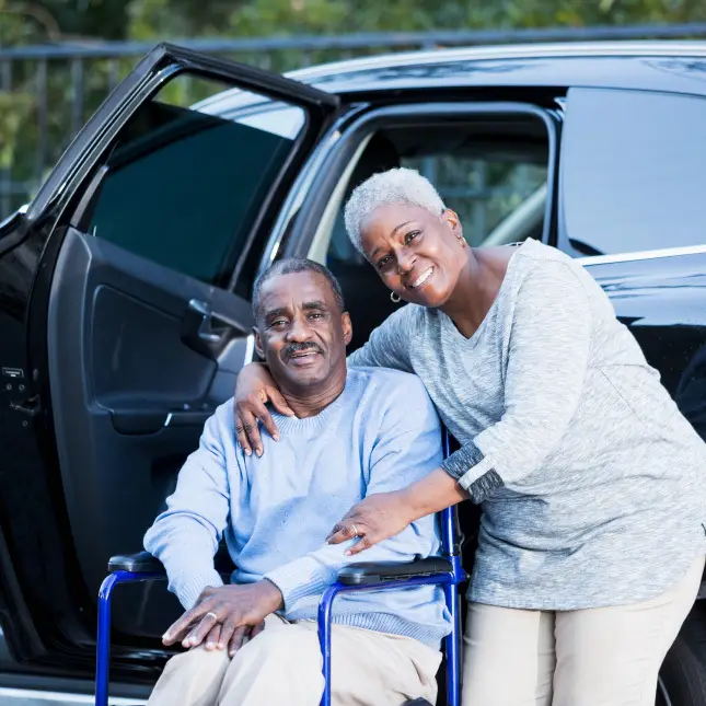 Driver wraps arm around member sitting a wheelchair while standing outside vehicle while smiling